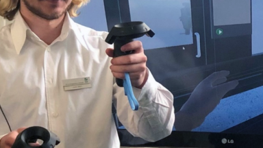 Nutzer mit VR-Headset und Controllern vor einem Bildschirm, auf dem die VR-Umgebung zu sehen ist.