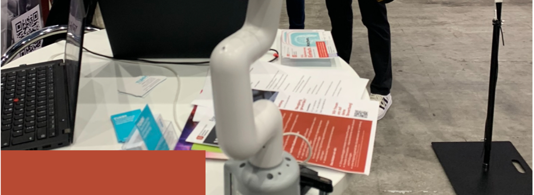 Ein weißer Roboterarm ist an einem Messestand auf einem Tisch befestigt. Der Roboterarm dient als interaktiver Blickfang und demonstriert automatisierte Bewegungsabläufe.
