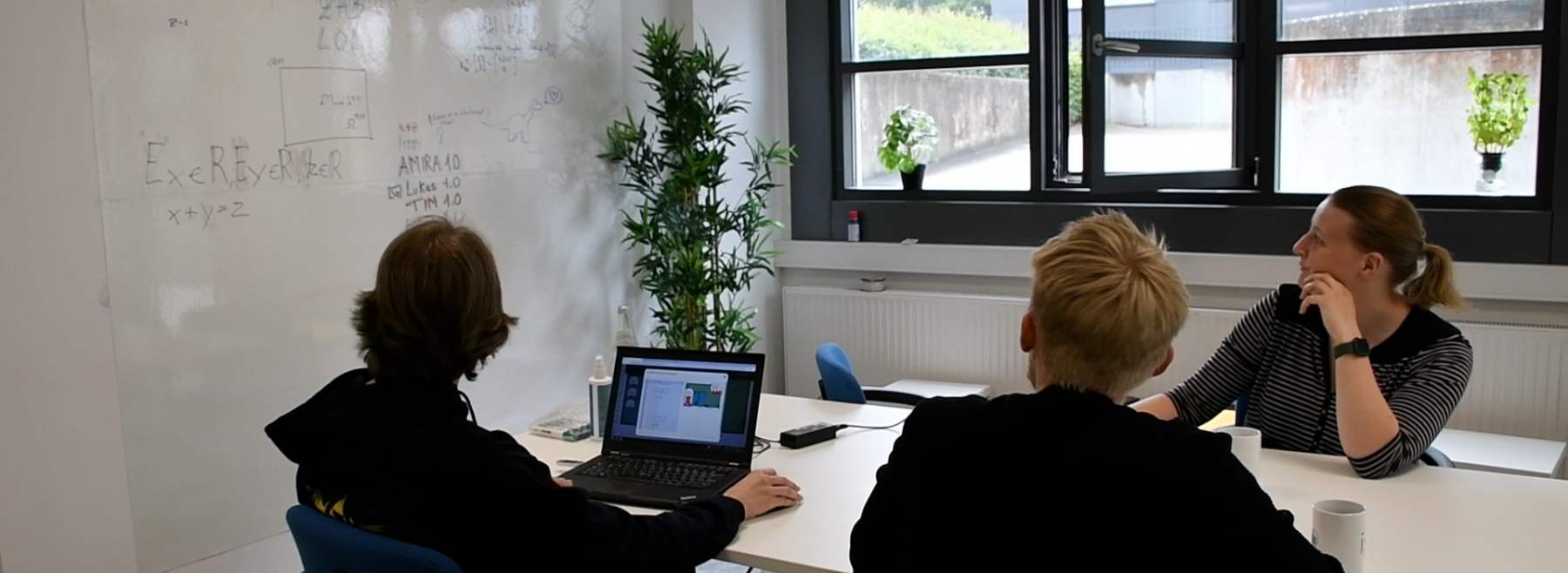 Drei Menschen an einem Tisch schauen auf ein beschriebenes Whiteboard