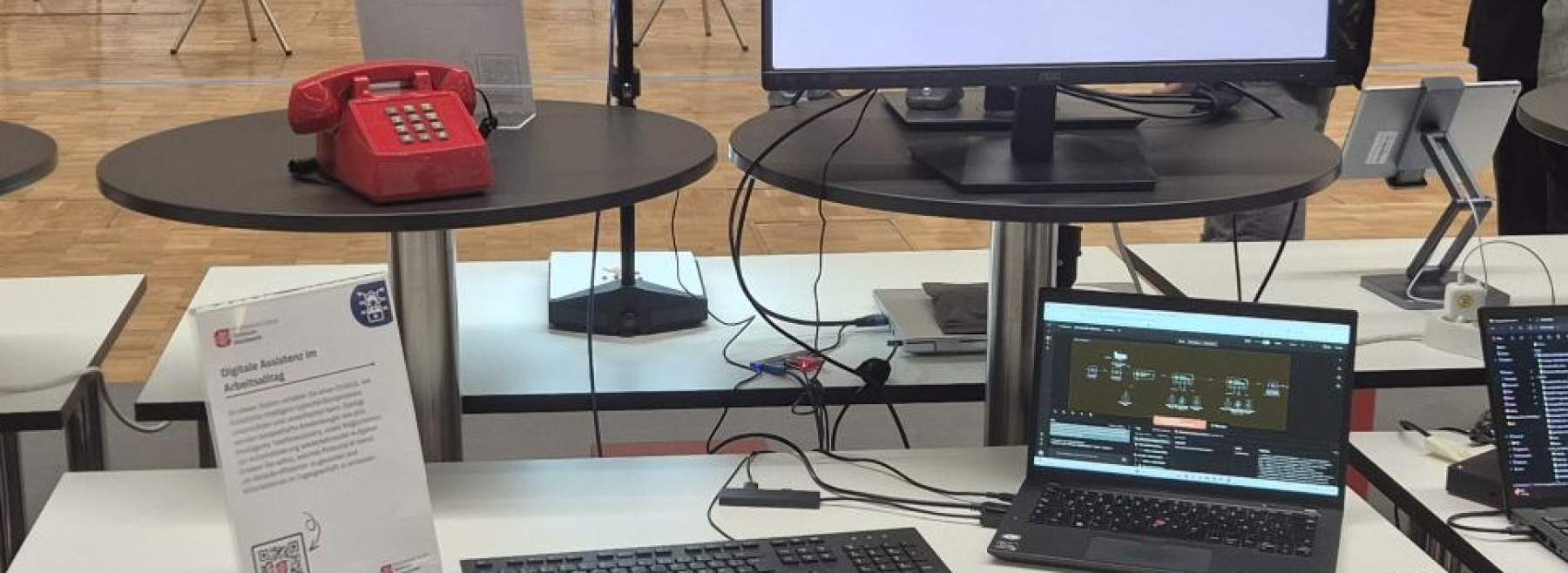 Physisiche Präsenz des Demonstrator KI im Büro. Aus dem Bild sind ein Bildschirm, Tastatur, Laptop, Maus und ein rotes Telefon.